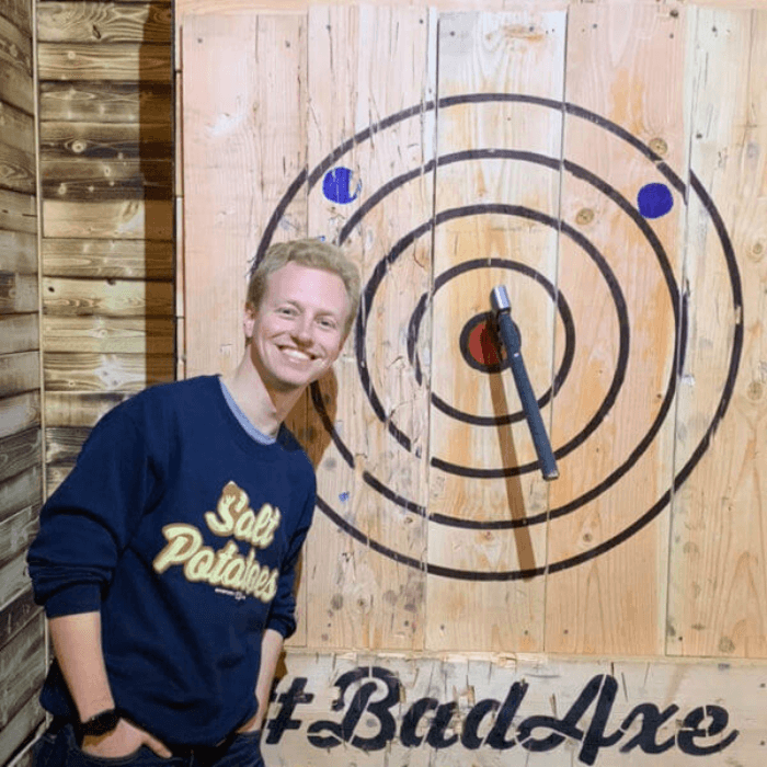 A man stands in front of a target proudly smiling at his well-thrown axe.