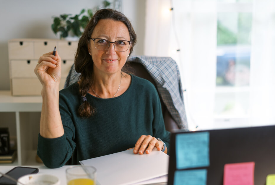 A woman with glasses sitting in a home office and writing in a notebook.