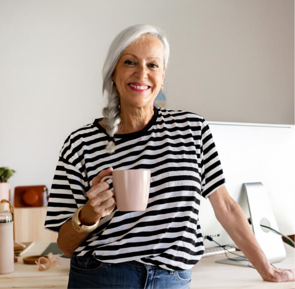 An older woman leaning on a desk holding a coffee cup.