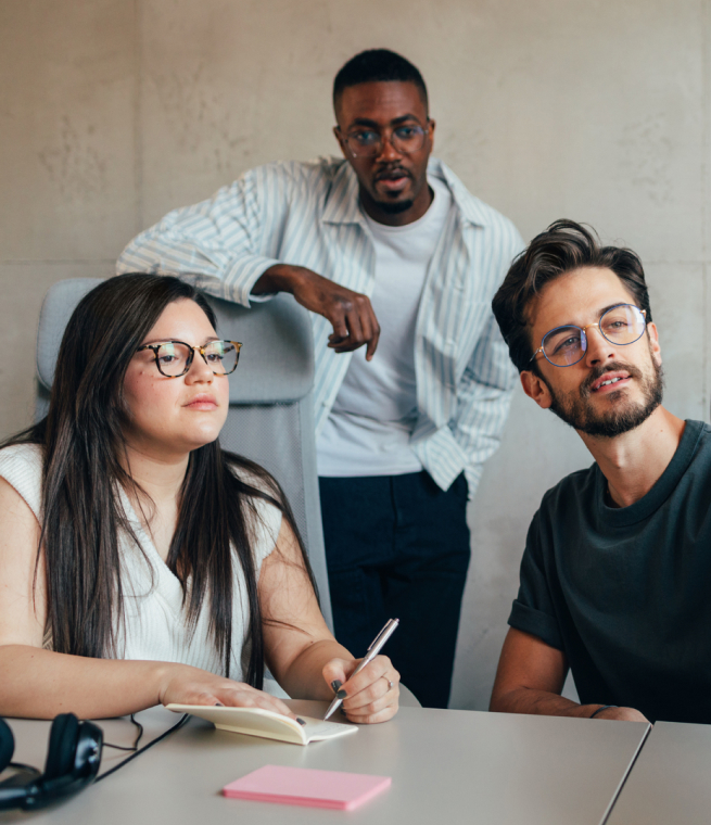 Three people, two men and a woman, collaborating and looking at a whiteboard in the distance.