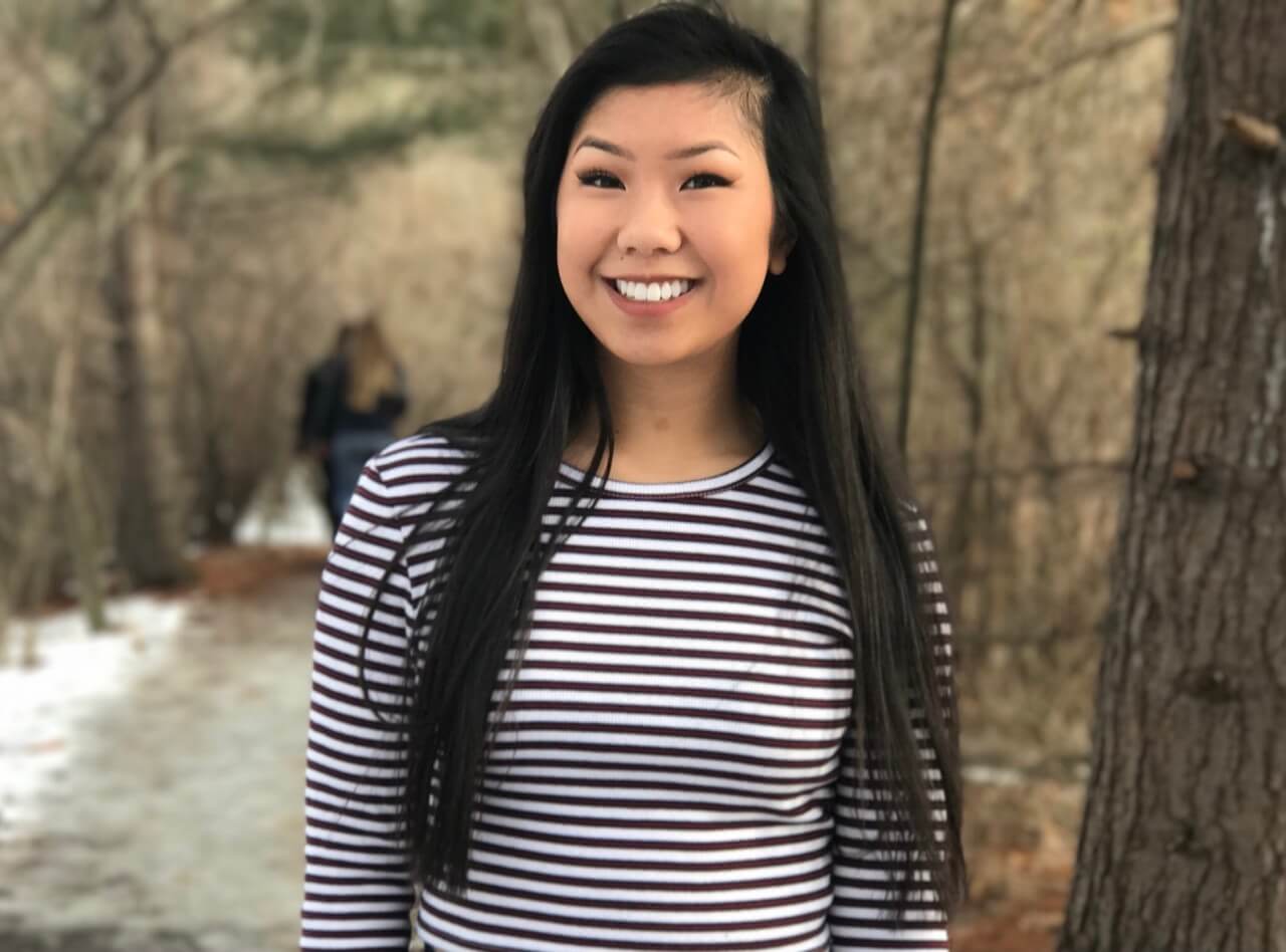 Cali Scahill stands smiling in front of some trees on a pretty hiking trail!