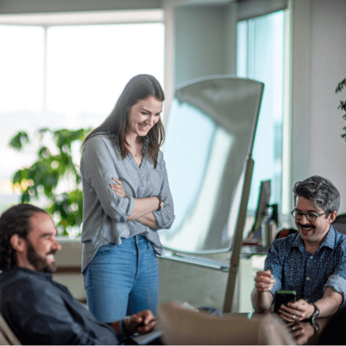 Woman stands above two seated men, laughing in an office.