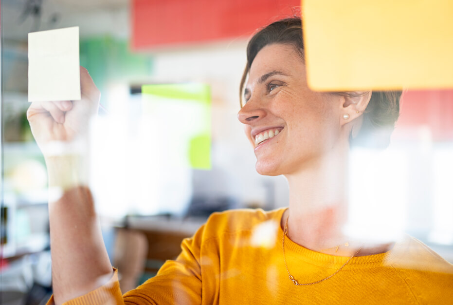 A woman in a yellow shirt smiling and writing on a sticky note.