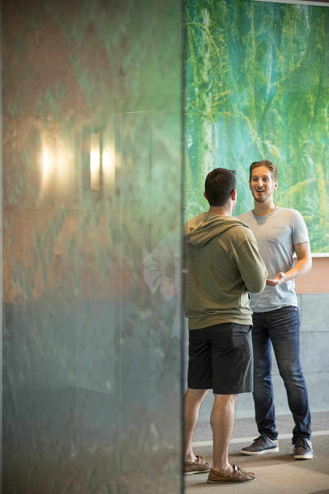 Two men stand and speak to each other in front of a green wall