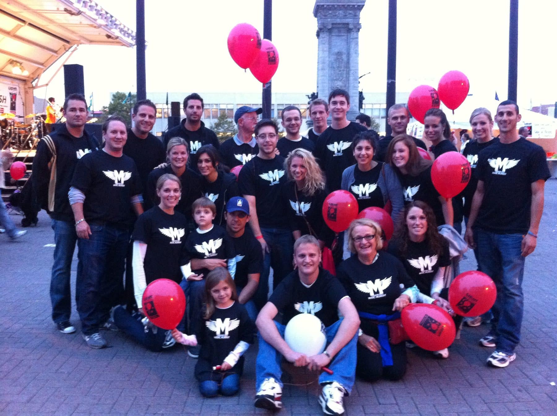 A large group of people pose, wearing matching shirts and carrying red balloons.