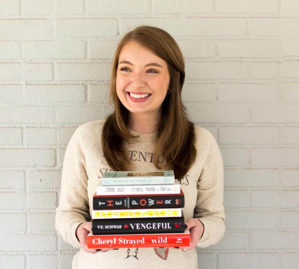 Alicia Riehle standing against a white brick background with a stack of books in her hands