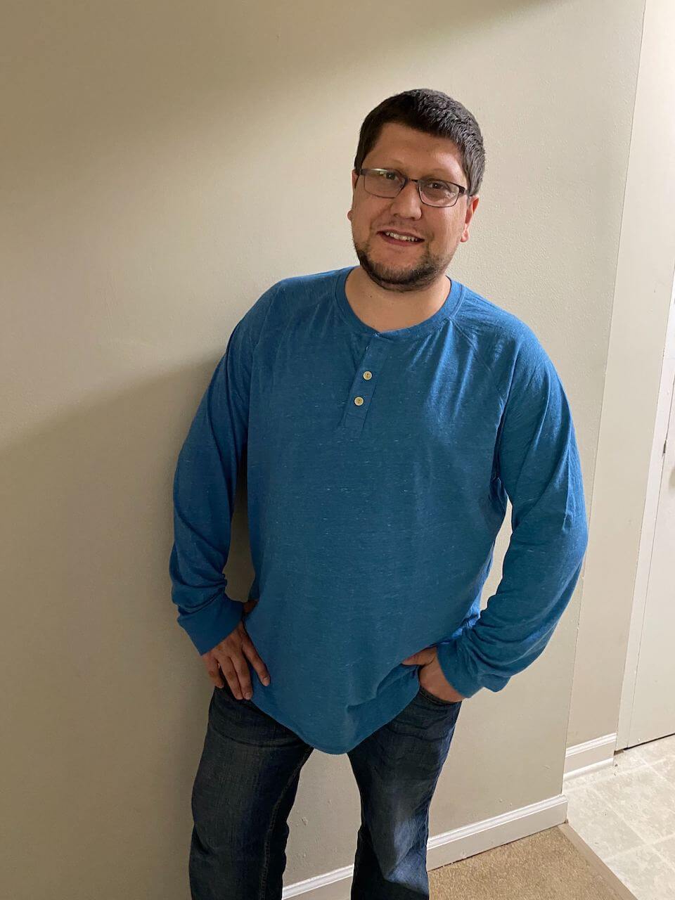 saj hoffman-hussein wearing blue shirt and jeans standing in front of a white wall.