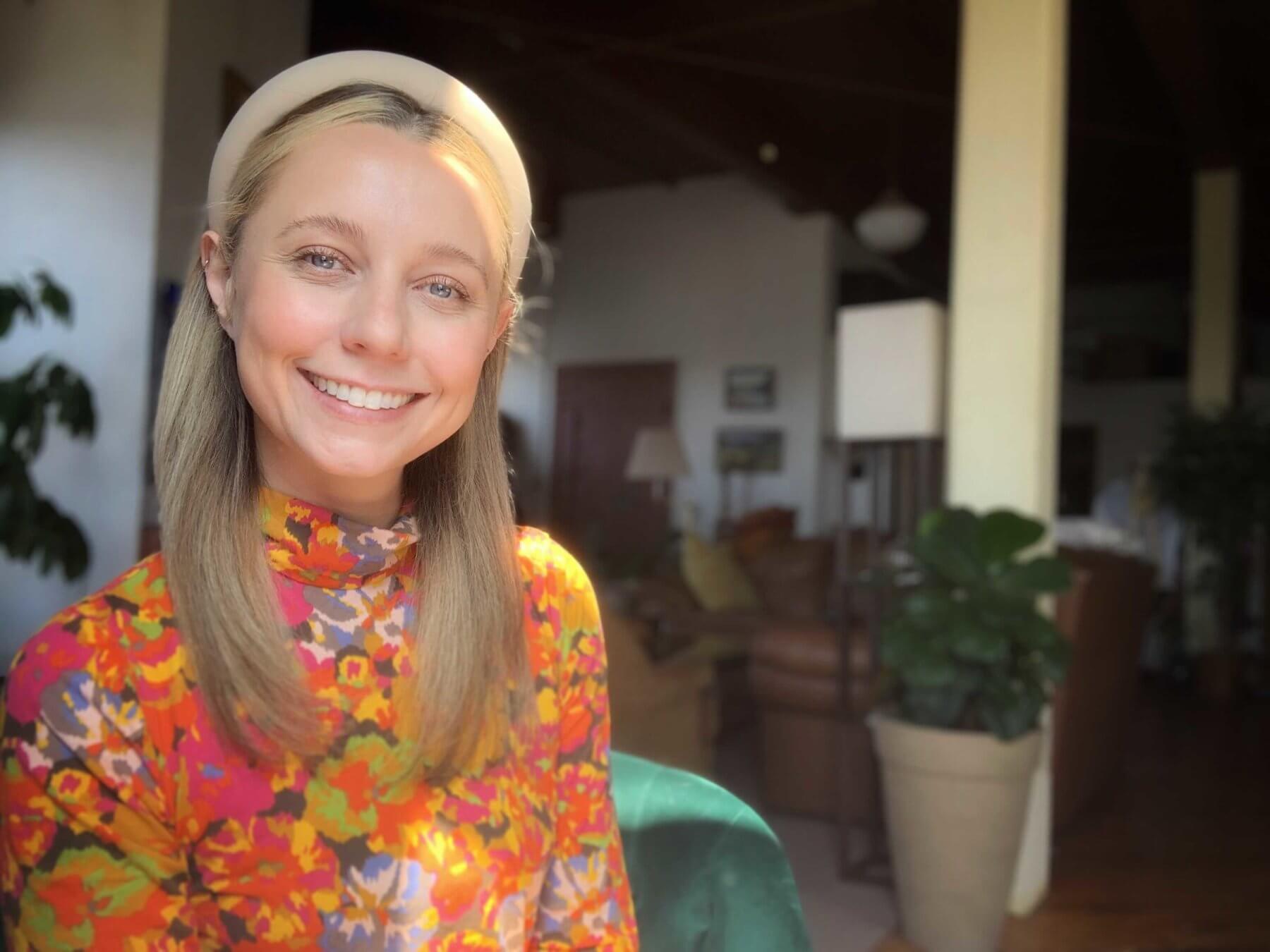 Kayla Mackie wears a floral orange top and smiles for the camera in her home office.
